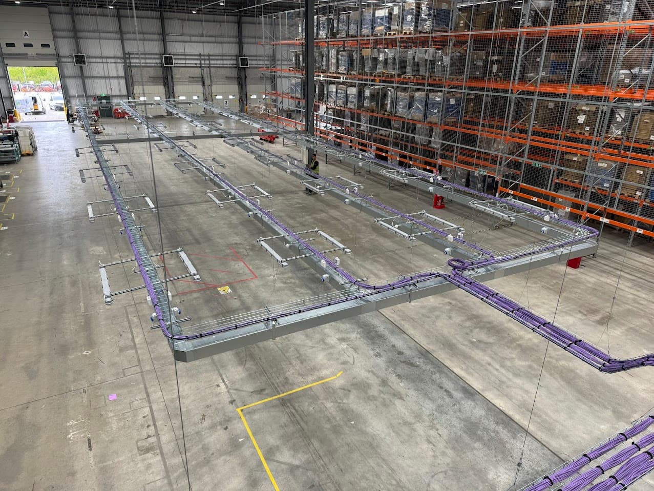 Steel cable containment installation carrying CAT6 data cabling above a packing area at Moonpig's Tamworth facility, with pallet racking and a loading door visible, and a member of the site team working nearby.