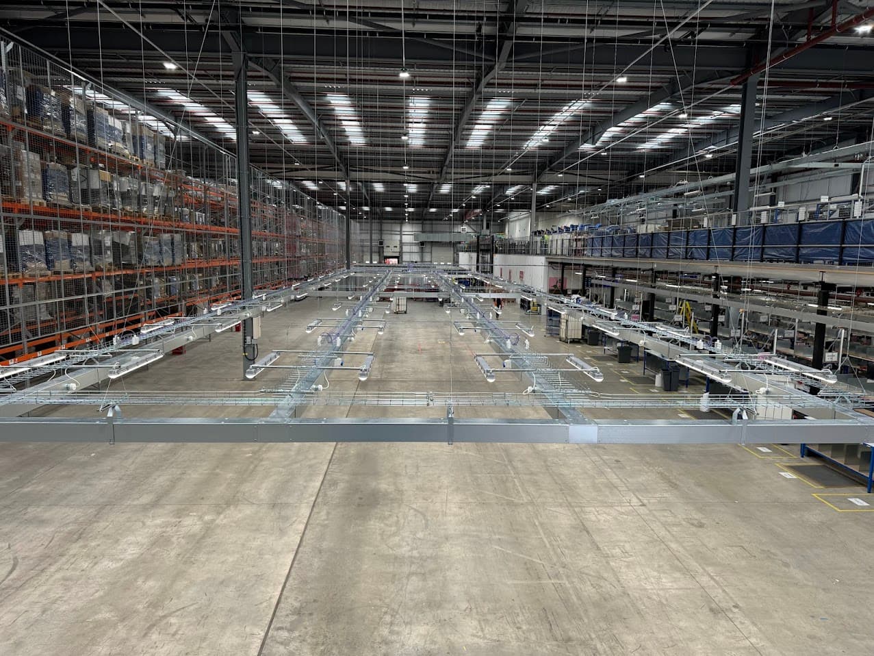 Elevated wide view of the completed electrical containment and packing bench infrastructure inside Moonpig's Tamworth distribution facility, showing steel containment runs and cable basket extending across the packing area, with pallet racking to one side and a mezzanine level beyond.