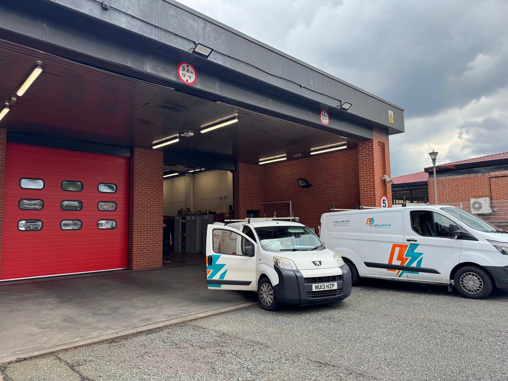 Exterior of a West Midlands Fire Service station showing the vehicle bay's roller shutter door, with two liveried RS Electrical vans parked at the entrance.