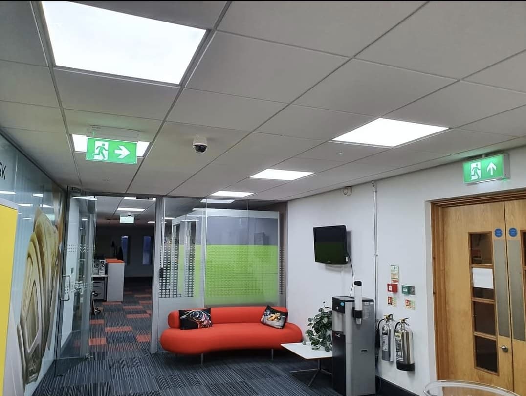 An open-plan office corridor with modern LED ceiling panels, illuminated exit signage and a seating area.
