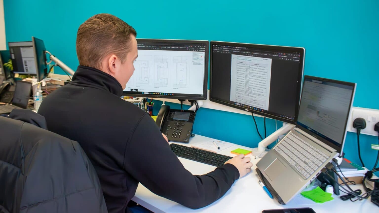 An RS Electrical team member at a multi-monitor desk reviewing drawings and documents while coordinating reactive electrical callouts.